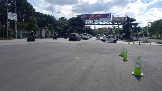 Wide road intersection in an urban area with several vehicles including cars and motorcycles moving through, green traffic cones placed along the edges, a large billboard structure overhead, trees and utility poles lining the sides under a partly cloudy sky