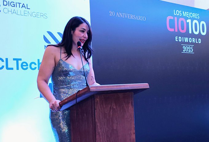 A woman with long dark hair wearing a sparkling silver dress stands at a wooden podium on a stage, speaking into a microphone held in her right hand. Behind her is a large blue backdrop with white text reading CIO1000 and LOS MEJORES CHALLENGERS, along with logos for EDIWORLD and CLTech, and a banner noting 20 ANIVERSARIO and 2025.