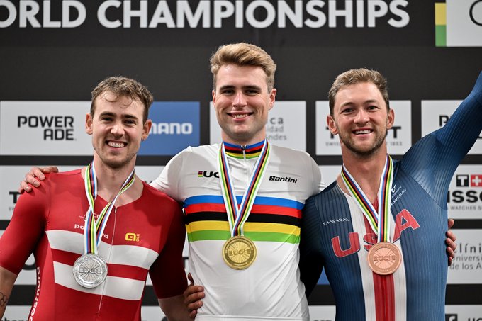 Three male athletes stand on a podium during the UCI Track Cycling World Championships. The central athlete wears a white suit with rainbow stripes and a gold medal around his neck, smiling and raising one arm. The left athlete in a red suit with white stripes holds a silver medal and smiles while placing a hand on the central athletes shoulder. The right athlete in a blue suit holds a bronze medal and smiles. Background includes event branding with World Championships text and sponsor logos like Powerade and Tissot.