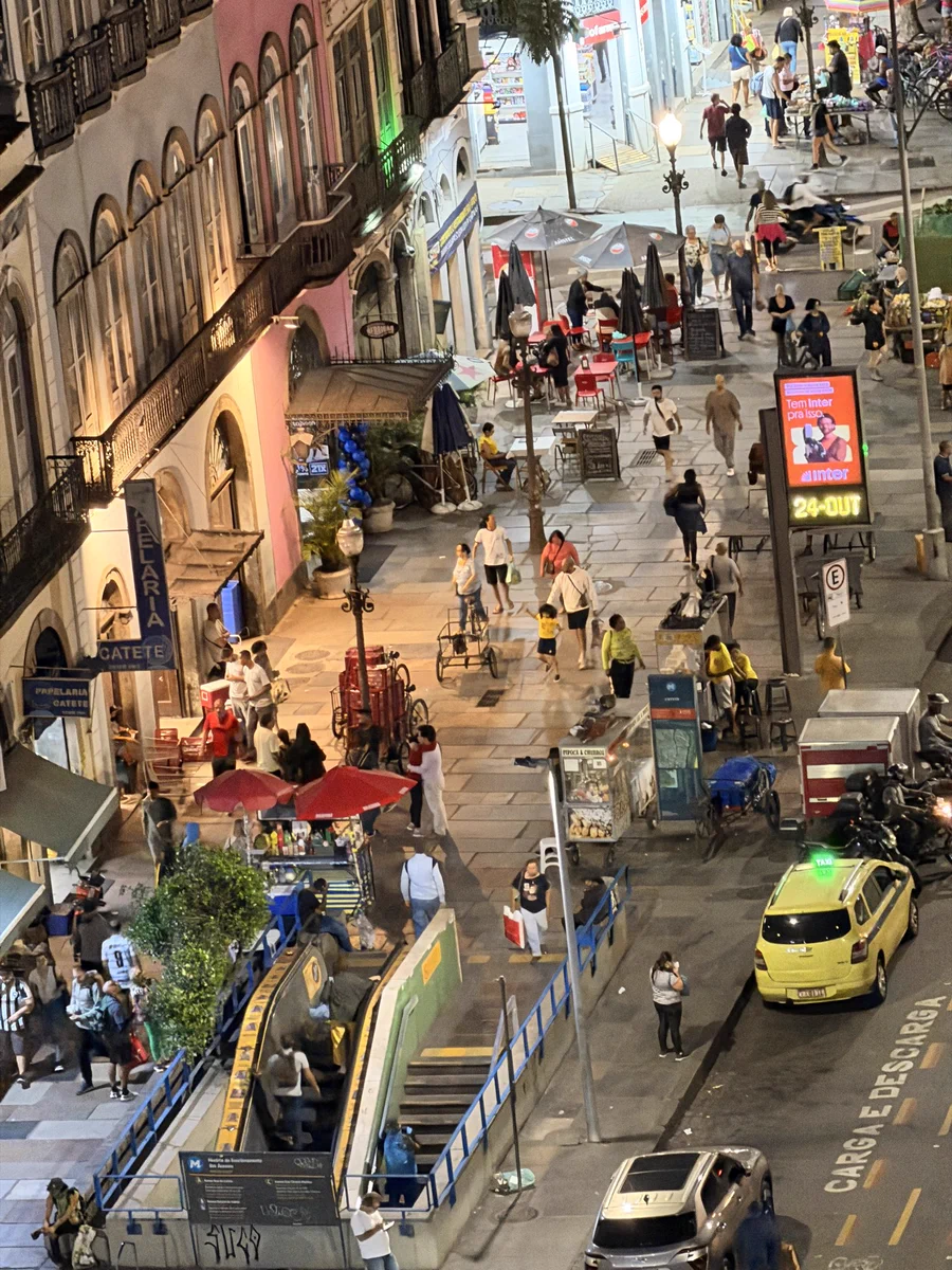 A fotografia mostra uma movimentada cena noturna na Rua do Catete, no Rio de Janeiro. A imagem é vibrante e cheia de vida, com pedestres caminhando pela calçada larga e iluminada, repleta de mesas de bares, carrinhos de comida de rua e toldos coloridos. À esquerda, fachadas antigas com varandas de ferro e paredes em tons de rosa e bege compõem o cenário típico da arquitetura carioca. O letreiro azul indica a entrada da estação de metrô Catete, por onde entram e saem várias pessoas. Na rua, um táxi amarelo e outros veículos passam devagar, enquanto ciclistas e entregadores ocupam o entorno. O ambiente urbano mistura o charme histórico dos prédios com a agitação contemporânea da cidade, criando um retrato autêntico da vida noturna carioca.