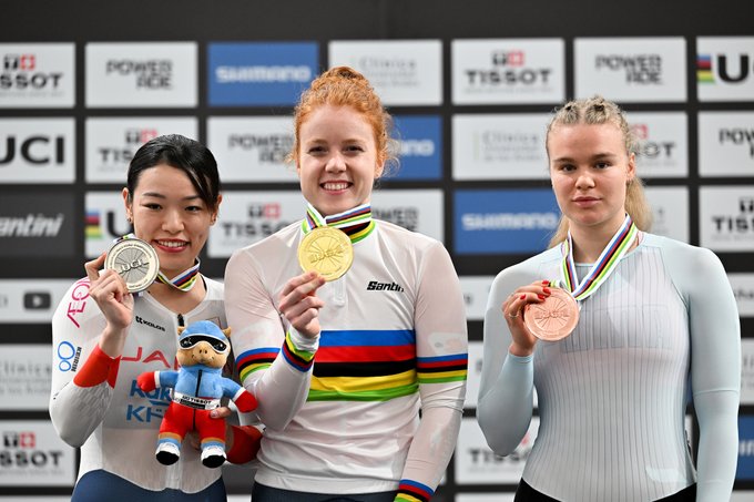 Three female athletes stand on a podium holding medals. Left athlete with red hair in Dutch kit holds gold medal and small plush toy. Center athlete with black hair in Japanese kit holds silver medal smiling. Right athlete with blonde hair in neutral kit holds bronze medal. Background features sponsor banners for Power2Cure, Samsung, Tissot, UCI, and event flags.