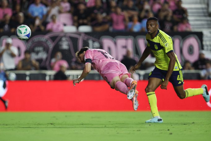 Lionel Messi in a pink kit dives low on green turf reaching for a green and white soccer ball mid-air while a player in yellow and black Adidas kit with dark skin and short hair extends a leg to challenge him the scene occurs on a stadium field with pink-shirted spectators in stands and a red barrier nearby under stadium lighting.