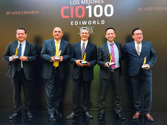 Five men in business suits and ties stand side by side holding golden trophies each with one hand while smiling at the camera in front of a dark backdrop featuring white text reading 20 Aniversario and bold red Los Mejores 100 CIO and CIO 100 logos the men have varied facial features including mustaches and glasses and wear polished shoes
