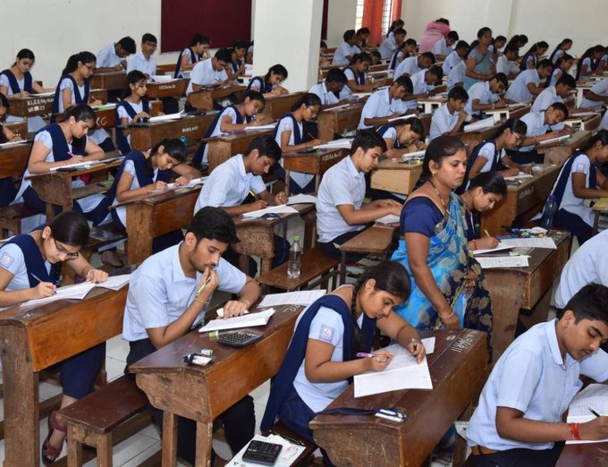 Classroom scene with rows of wooden desks and benches occupied by male and female students in blue and white school uniforms sitting attentively. Students are writing on papers using pens and pencils with some using calculators. A female teacher in a blue sari stands near the front supervising. Blackboard on the wall and windows with curtains in the background.