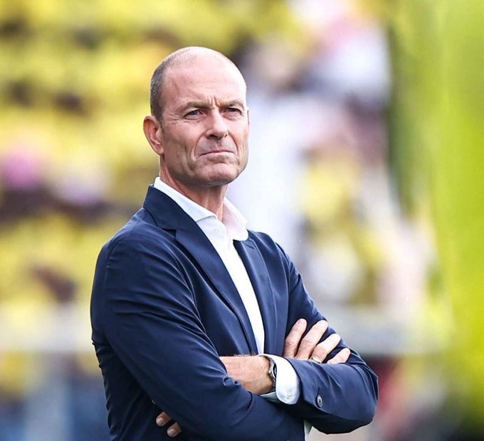 A bald man with a serious expression wearing a dark blue suit white shirt and watch stands with arms crossed in front of a blurred yellow and green stadium background with spectators visible.