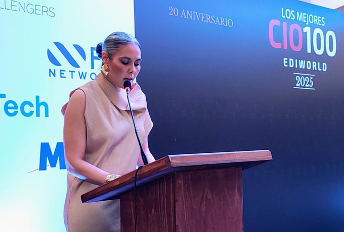 A woman with light hair in a ponytail wearing a sleeveless beige dress and gold earrings stands at a wooden podium holding a microphone. Behind her is a large screen displaying text LOS MEJORES CIO1000 EDiworld 20 ANIVERSARIO TECH NETWORKS and logos including HCLTech and others. The setting appears to be an indoor event stage with blue and white lighting.
