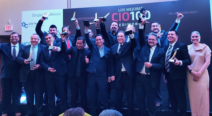 Group of professionals in suits and formal wear standing on a stage holding trophies raised in celebration. Backdrop displays banners for Grupo Mexico and CIO100 20th anniversary event. Setting appears to be an indoor conference or award ceremony with lighting and signage visible.