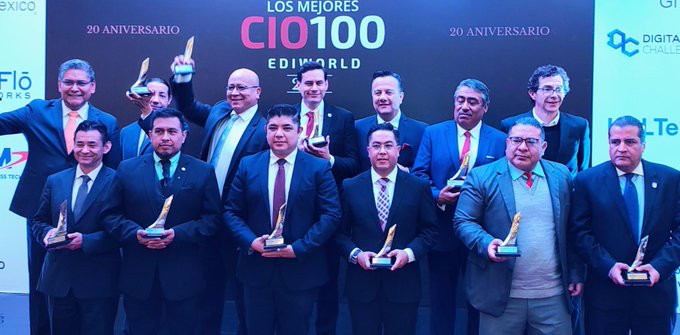 Group of about 12 men in business suits standing on a stage holding trophies, smiling and posing for the camera, with a large banner behind them reading 20 Aniversario CIOs Mejores100 20 Aniversario and logos including CIO EDIWORLD, Digital Future, and sponsor names like Huawei and Microsoft visible on the backdrop and podium.