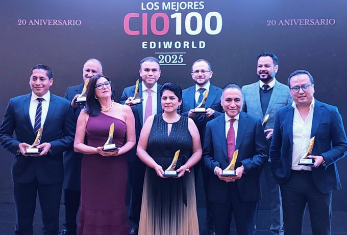 Group of ten professionals, eight men and two women, standing on a stage holding golden trophies. They wear formal business attire including suits, ties, dresses, and blazers. Backdrop features text LOS MEJORES CIO100 20 ANIVERSARIO EDIWORLD 2025 in white and red lettering.