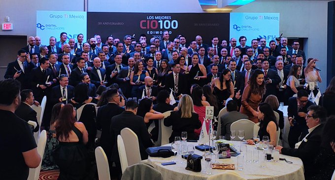 Group of professionals in formal attire stands on stage behind podiums during event with banners displaying Grado MX and CIO 100 logos on screens. Front table features glasses wine bottles and awards. Attendees include men in suits and women in dresses smiling and posing together in conference setting.