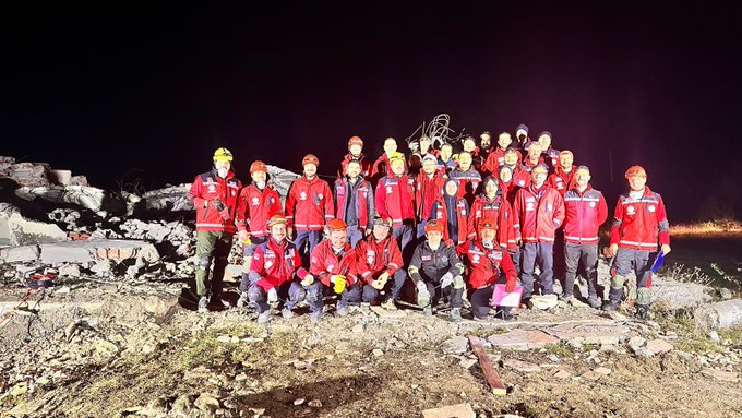 Group of rescuers in red uniforms and yellow helmets standing together on rocky terrain at night during a disaster drill in Kastamonu. Another image shows a rescuer in red gear lying on the ground near a manhole cover with team members surrounding the scene. Third image depicts rescuers in red outfits working near a white ambulance marked with Turkish Ministry of National Education and MEB AKUB logos on a debris-strewn area at night.