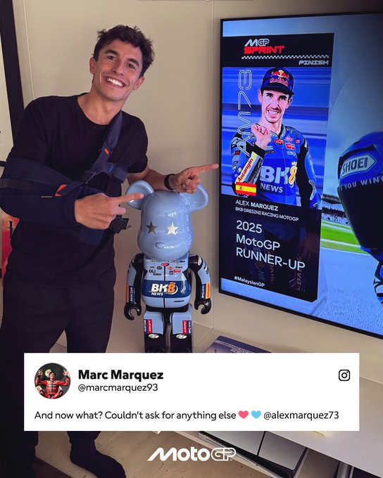 Marc Marquez stands smiling in black clothing with a camera strap pointing at a large blue Bearbrick toy figure of Alex Marquez in red racing suit and helmet next to a wall-mounted poster showing Alex Marquez on a motorcycle with 2025 MotoGP branding and sponsor logos including Red Bull and Hublot.