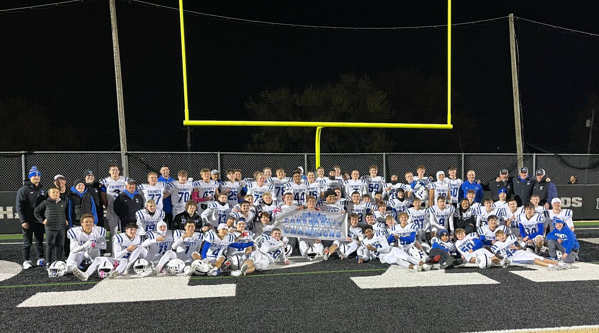 Group of high school football players and coaches in white and blue uniforms pose together at night on a field in front of yellow goalposts and a chain-link fence. They hold a blue banner with white text reading CONFERENCE CHAMPIONS. Players kneel or stand in formation, some wearing helmets, under stadium lights with a dark sky above. The field has white yard lines visible on the grass.