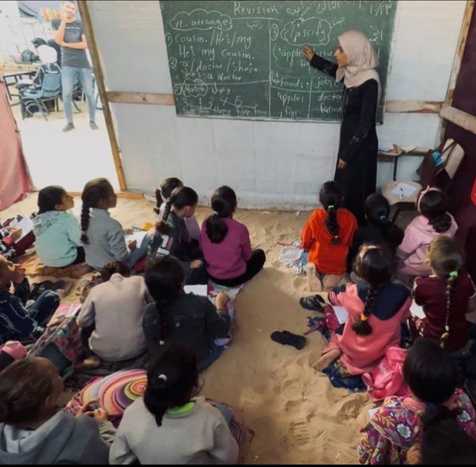 Teacher wearing a black hijab and abaya stands in front of a blackboard covered in handwritten English notes including lists like My Files, Applications, and other lesson elements. Several young girls with colorful headscarves and casual clothing sit cross-legged on a sandy floor, holding notebooks and looking attentive. Classroom has simple wooden desks, chairs, and an open door revealing outdoor light. Setting appears makeshift with dirt ground inside.