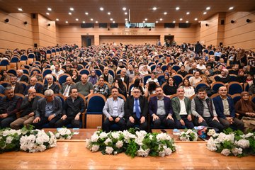 First image shows a large group of men in suits and formal attire standing on a wooden stage with blue backdrop banners displaying text like BEUN and event names surrounded by green plants and white flowers at the edges. Second image depicts a crowded auditorium filled with audience members seated in blue chairs facing a stage with wooden floor and floral decorations. Third image captures three male performers on stage one with a beard holding a microphone another in traditional vest gesturing with hand on chest and the third standing nearby with backdrop banners reading SAMİL YOLU and CİĞER. Fourth image features three actors in a scene one with beard in white shirt and vest another in black shirt touching shoulder and the third in black top sitting at a table with chessboard and chairs against backdrops with Turkish text like BİR İNSAN KALBİMDE BİR and VEFA CİĞER.