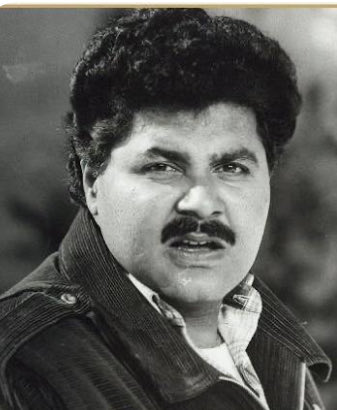 Black-and-white portrait of Satish Shah, a man with curly black hair, thick mustache, intense expression, wearing a collared shirt under a dark jacket with lapels.