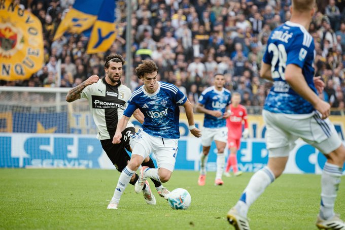 Football players in blue and white uniforms compete on a green field during a match with a crowd in the background holding blue flags and banners including one with a yellow emblem near the goal area while another player in black and white advances with the ball