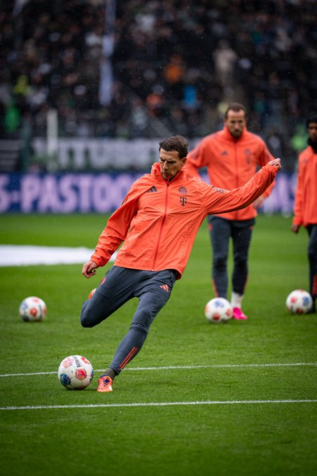 A soccer player in an orange Adidas jacket and gray pants kicks a white soccer ball on a green field during training with other players in similar orange jackets standing nearby and several soccer balls scattered around the pitch under stadium seating in the background
