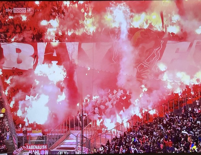 Vibrant stadium scene filled with red smoke from pyrotechnics creating a dramatic atmosphere, large crowd of fans in stands holding a massive banner with white letters spelling BAYERN on red background, additional red and white smoke billowing around, metal railings and stadium structure visible in foreground, capturing excitement of FC Bayern Munich football event.