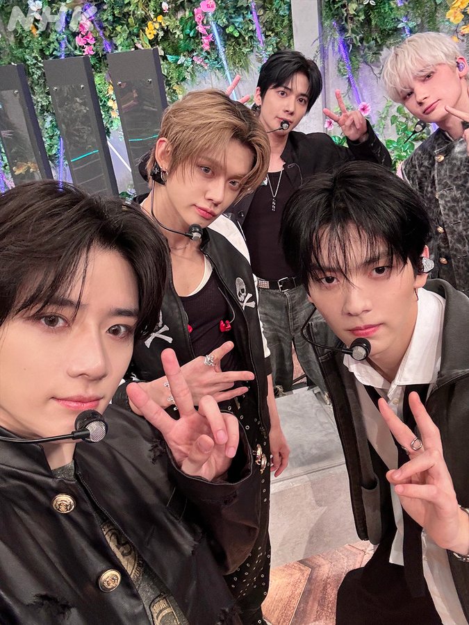 Five young men with varied hairstyles including black, blonde, and silver, dressed in black and colorful outfits with accessories like chains and belts, standing on a stage surrounded by large green leafy plants and purple lights, holding microphones, making peace signs with hands, and smiling at the camera.