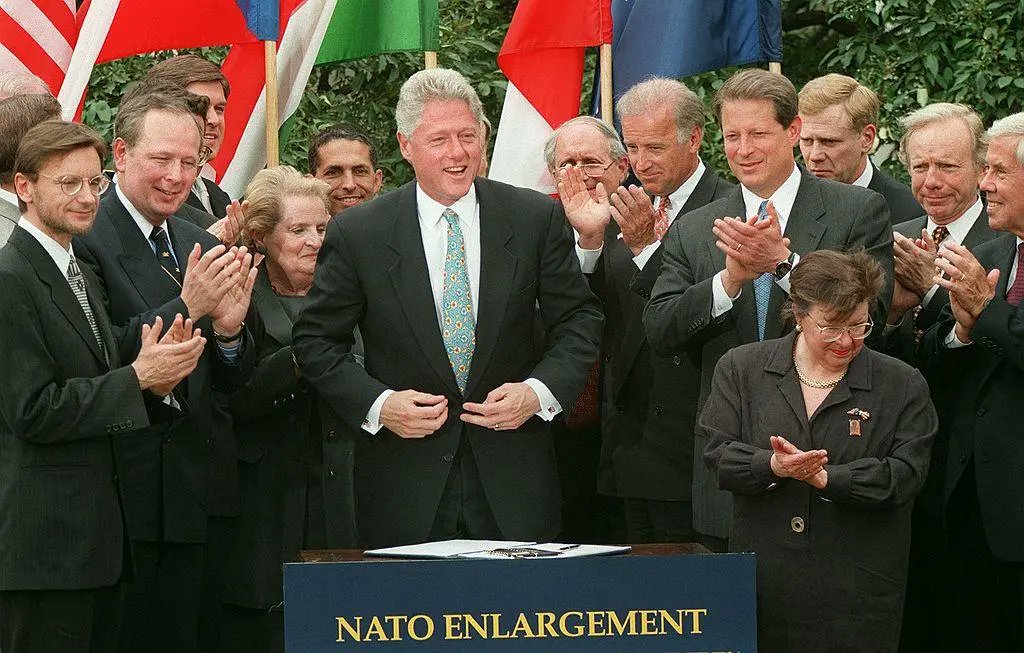 Group of world leaders including Bill Clinton standing and clapping in formal suits and dresses at a podium labeled NATO Enlargement, with flags of various countries such as the United States, France, Germany, Italy, and others in the background, set outdoors with greenery.