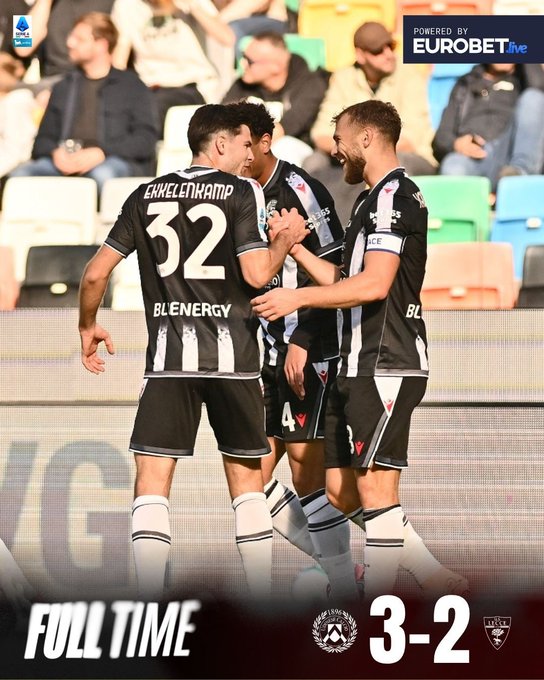 Three male soccer players in black and white Udinese uniforms with number 32 on one jersey stand on a green field facing away from the camera, arms linked in celebration, surrounded by stadium seats with spectators, yellow and green elements visible, full time graphic shows 3-2 score between Udinese and Lecce crests, Eurobet sponsor logo present.