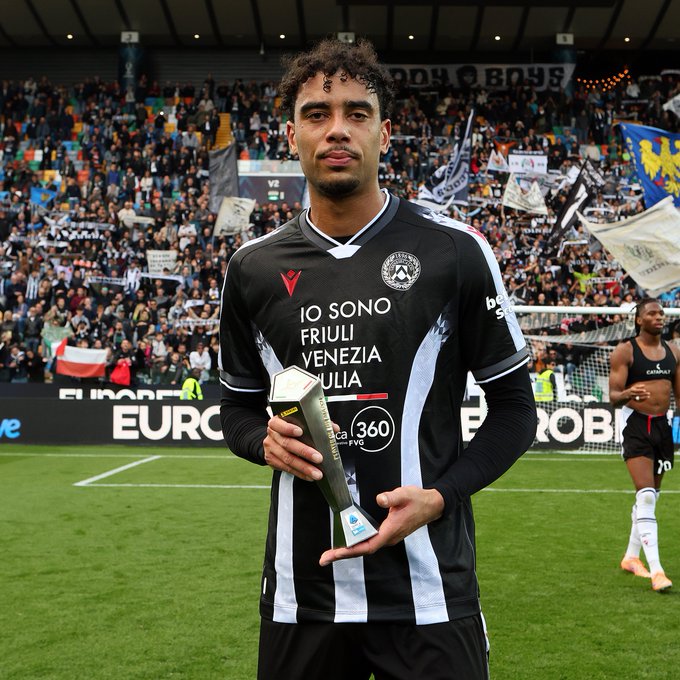 Arthur Atta, a young Black male athlete with curly hair, stands on a soccer field holding a silver trophy engraved with Panini branding, wearing a black and white Udinese team jersey with sponsor logos including Io Sono Venezia and V, black pants, and long sleeves, surrounded by stadium seating filled with fans waving blue and white flags, another player in similar kit visible nearby, green field and goalpost in background.