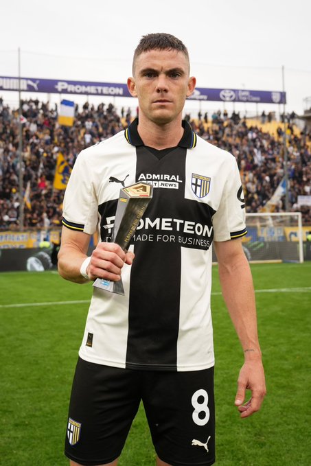 A young man with short dark hair stands on a green soccer field holding a silver trophy with both hands. He wears a white and black vertically striped short-sleeved jersey with gold accents, the number 8 on the front, and black shorts. The jersey features sponsor logos including Puma, Admiral, and Made for Business. In the background, a stadium with stands filled with spectators, advertising banners for Prometeon and Toyota, and a blue sky.