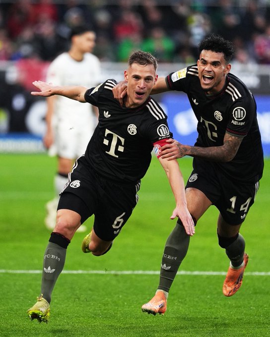 Two Bayern Munich players celebrate a goal on a soccer field. Harry Kane, wearing jersey number 16, has short brown hair, a focused expression, and raises both arms while in a black Adidas kit with white FC Bayern logo, T-Mobile sponsor patch, and gray socks ending in green and orange cleats. Alphonso Davies, in jersey number 19, has dark curly hair, a wide smile, and an arm around Kane, showing a tattoo on his left arm; he wears similar black kit with gray socks and orange cleats. Stadium seating with blurred spectators and green pitch in background.