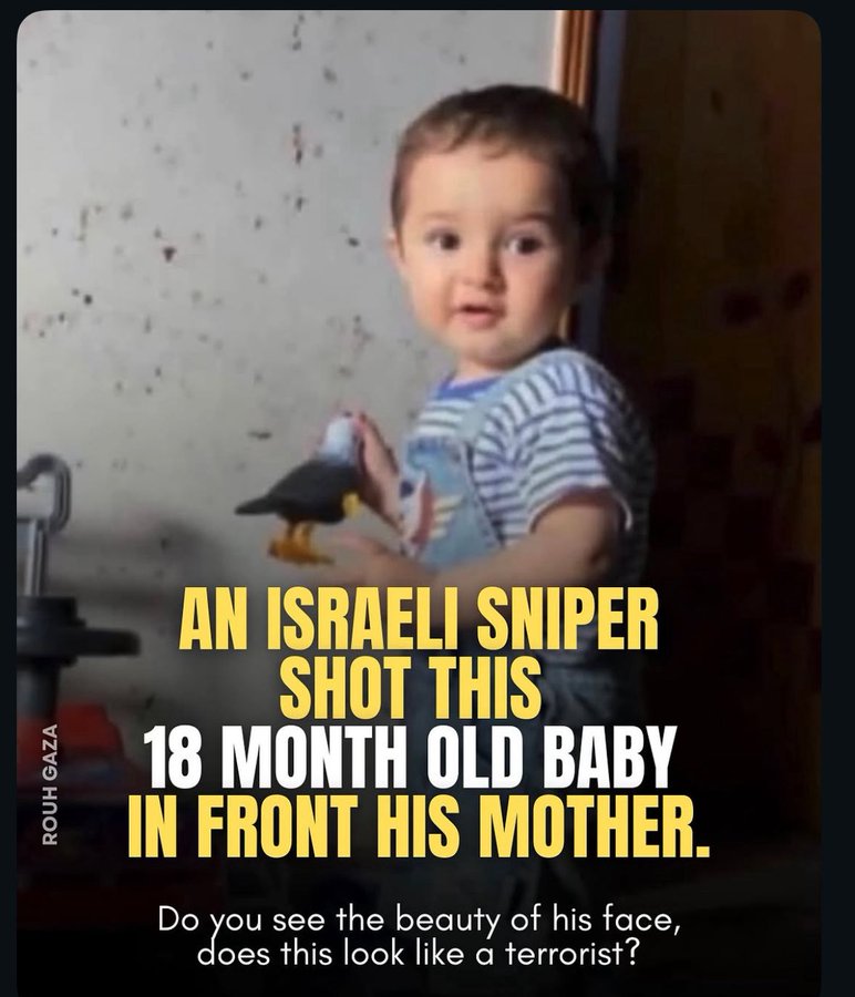 A young boy with light brown hair and wide eyes smiles while holding a small black bird toy with yellow beak in his hands. He wears a light blue and white striped short-sleeved outfit and stands in front of a plain beige wall. The image has yellow overlaid text reading AN ISRAELI SNIPER 18 MONTH OLD BABY IN FRONT HIS MOTHER. DO YOU SEE THE BEAUTY OF HIS FACE DOES THIS LOOK LIKE A TERRORIST.