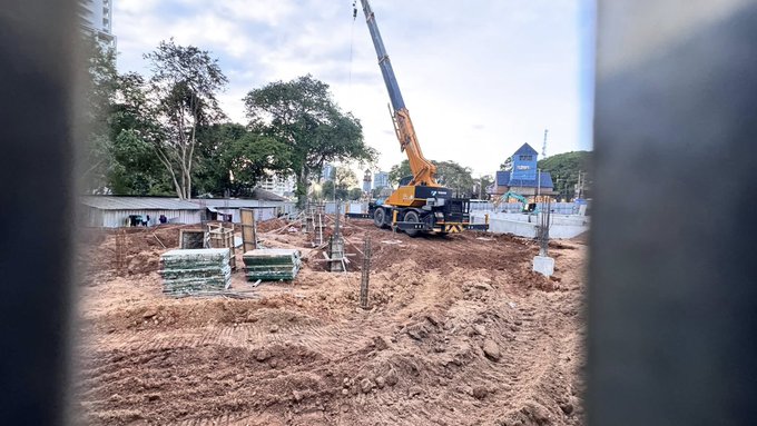 Orange crane with extended arm positioned over dirt ground at construction site surrounded by stacked wooden pallets metal beams and concrete pillars partial blue building structure visible in background tall green trees frame the scene under partly cloudy sky no people or vehicles present