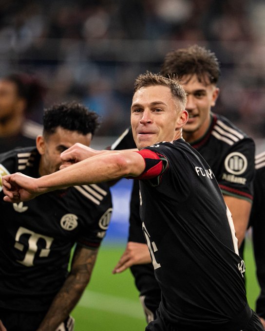 Players in black jerseys with red accents and sponsor logos stand on a green field in a stadium with blurred crowd background, one central player with short hair raises his arm holding a yellow object while smiling, others extend arms in celebration, tattoos visible on one arm.