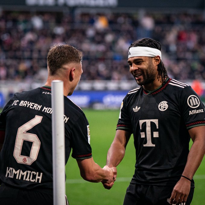 Two male soccer players stand on green field facing each other extending hands for handshake one with short brown hair wearing Bayern Munich jersey number 6 other with long dark hair tied in white headband smiling broadly both in black jerseys with green accents and sponsor logos including Allianz and Adidas stadium seating and crowd visible in blurred background upright pole between them FC Bayern Munich text on jersey backs