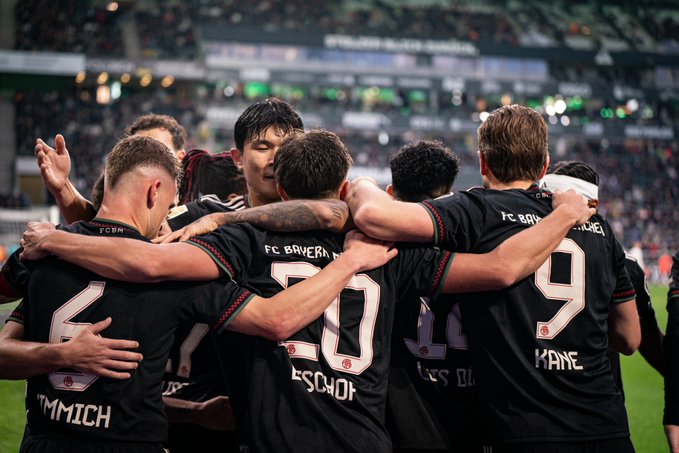 Group of Bayern Munich players in black red and white jerseys with numbers like 9 20 and names such as Mane and Gnabry stand closely embracing each other arms around shoulders on green soccer field under stadium floodlights with blurred green seats and audience visible in background players show joyful expressions with raised hands and smiles