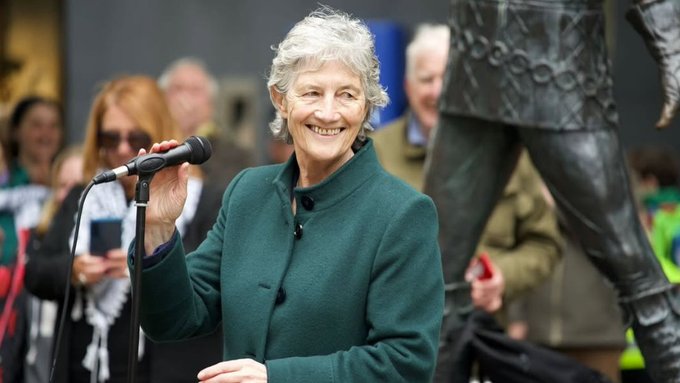 Catherine Connolly, an older woman with short gray hair wearing a green coat, smiles while holding a microphone and speaking at an outdoor public event. A crowd of people, including men and women in casual attire, stands behind her taking photos with phones. A large statue of a figure in draped clothing is visible in the background next to her. The setting appears to be a sunny urban square with some green elements.