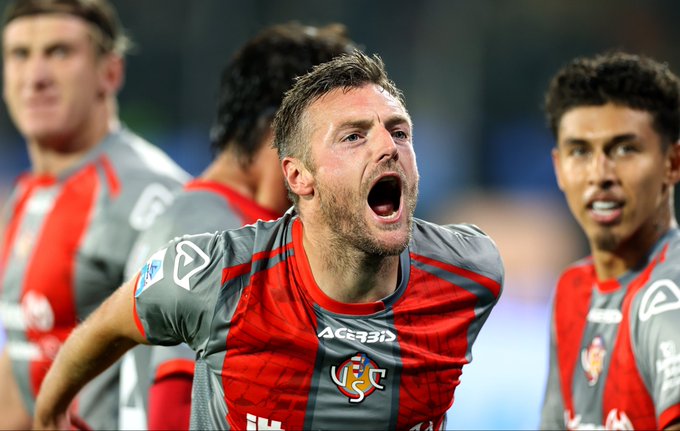 Group of male soccer players in red and gray uniforms with ACERBIS sponsor logos and U.S. Cremonese emblems stand on a lit stadium field, one central player with short brown hair shouts with mouth wide open and arms extended, another with dark hair smiles nearby, background shows blurred teammates and blue seating.