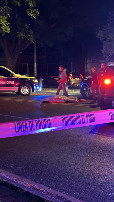 Nighttime street scene with police vehicles including a yellow truck with lights on and a black car. Blue and red emergency lights illuminate the area. A body covered by a red sheet lies on the ground near the vehicles. Pink police tape marked Linea de Policia Prohibido el Paso stretches across the road blocking access. Trees and a fence are visible in the background.