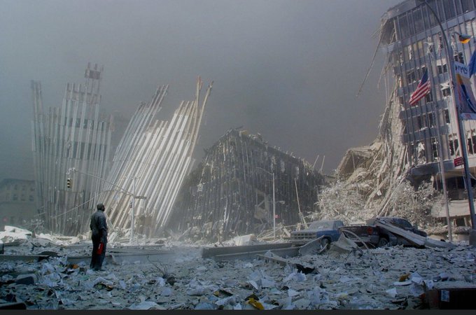 A historical photograph depicting the aftermath of the 9/11 attacks at Ground Zero in New York City, showing the skeletal remains of the collapsed World Trade Center towers amid smoke and debris, with scattered papers and rubble on the ground; a lone figure in work attire, possibly a firefighter or rescue worker, stands in the foreground facing the destruction, under a hazy, overcast sky.