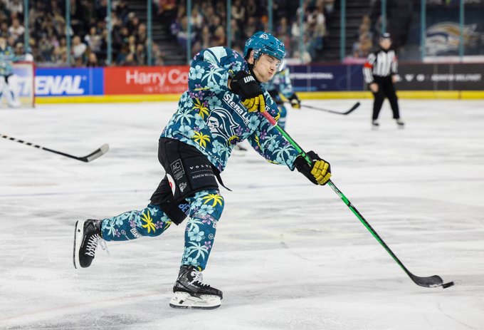 A hockey player from the Belfast Giants team wearing a bright turquoise helmet blue jersey with floral patterns black pants and yellow gloves skates dynamically on the ice while gripping a green stick and pursuing the puck near the goal area. Teammates in matching uniforms are visible in the background along with a referee in black and white stripes. The rink features advertising banners including one for Enal and Harry Corry with a crowd of spectators seated behind glass panels.