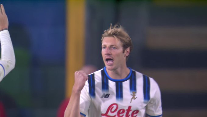 A male soccer player with blonde hair wearing a white and blue striped Atalanta jersey with NB logo and sponsor patch celebrates by raising his arms and shouting, standing on a green field with blurred stadium seating and another player in the background.