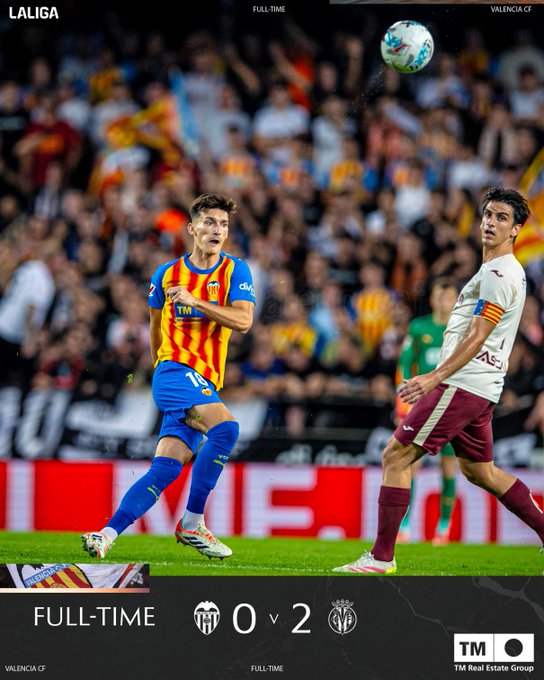 Soccer match action on a green field with a player in orange and black striped jersey number 7 kicking a ball towards goal defended by a player in white and burgundy kit number 24 both wearing white shorts and cleats stadium crowd in background upper text overlay reads LaLiga Full Time lower section shows score TM 0 2 with team logos Valencia CF and Villarreal CF and Full Time text watermarks from LaLiga and TM Group visible