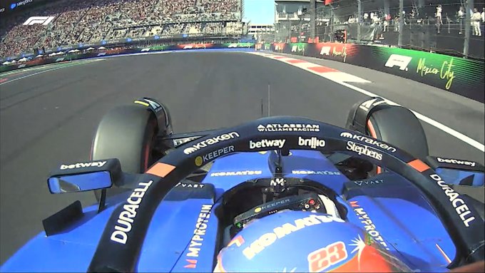 First-person view from inside a blue Formula 1 race car cockpit speeding along a straight track at a circuit with green barriers, sponsor logos on the steering wheel including Betway and Pirelli, front wheels visible, number 23 on the side, Mexico City branding on barriers, crowd in background stands under sunny sky.