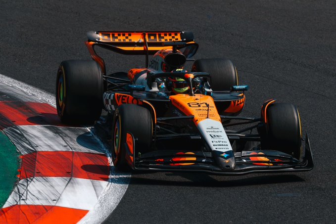 Orange and black McLaren Formula 1 race car with sponsor logos including Veloster and number 81 navigates a curved asphalt track section marked by green and white curbs and barriers during a race session with a driver wearing a helmet visible in the cockpit
