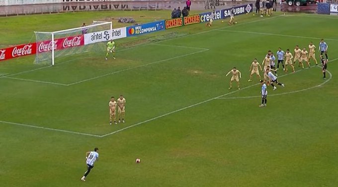 Green soccer field with goalpost and net on the left side surrounded by advertising banners for brands like Coca-Cola and Claro. Several players in yellow and black uniforms positioned defensively near the goal. One player in a blue and yellow uniform numbered 1 running towards the ball near the penalty area. Another player in blue and yellow kicking the ball. Spectators visible in the background stands.