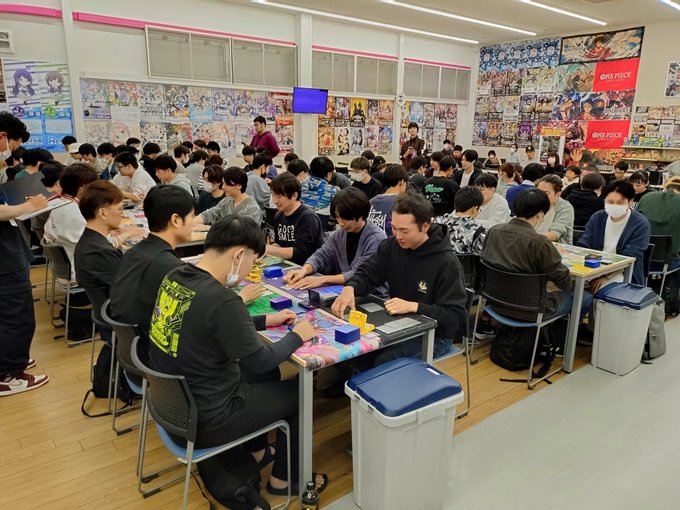 Interior of a trading card store with pink and white walls covered in posters of anime characters and promotional banners. Numerous young men and some women seated at long tables playing Pokemon card games, shuffling decks, and discussing moves. Tables hold colorful card sleeves, playmats with Pokemon artwork, and small cups. Store shelves in background display card packs and merchandise. Bright fluorescent lighting illuminates the spacious room with blue trash bins nearby.
