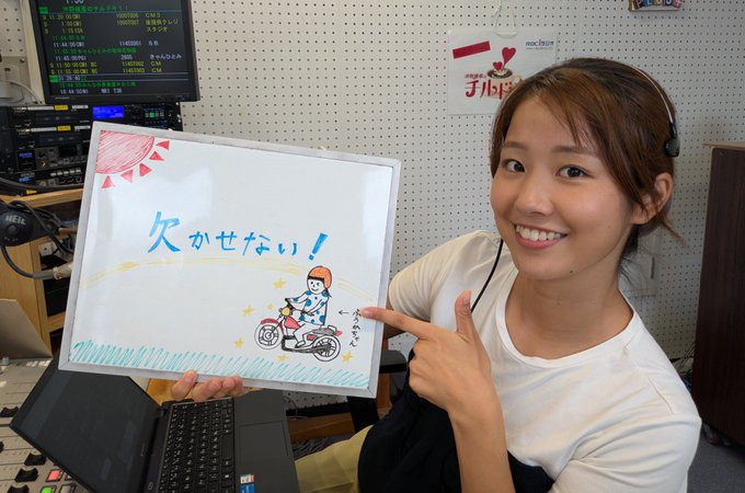 A young woman with brown hair wearing a white shirt and headset sits in a radio studio with monitors and equipment behind her, holding a whiteboard that has a hand-drawn sun, the Japanese text for summer vacation, a cartoon boy on a bicycle, and she points to it with a smile.