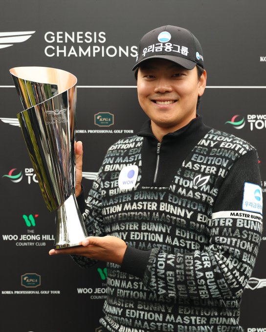 A man identified as Junghwan Lee stands smiling while holding a large silver trophy with his right hand. He wears a black cap with Genesis logo and a black zip-up jacket covered in white text reading Master Bunny Edition repeated across it. The background features black panels with white text for Genesis Championship in Korean and English, DP World Tour logo, and Woo Jeong Hill Country Club branding.