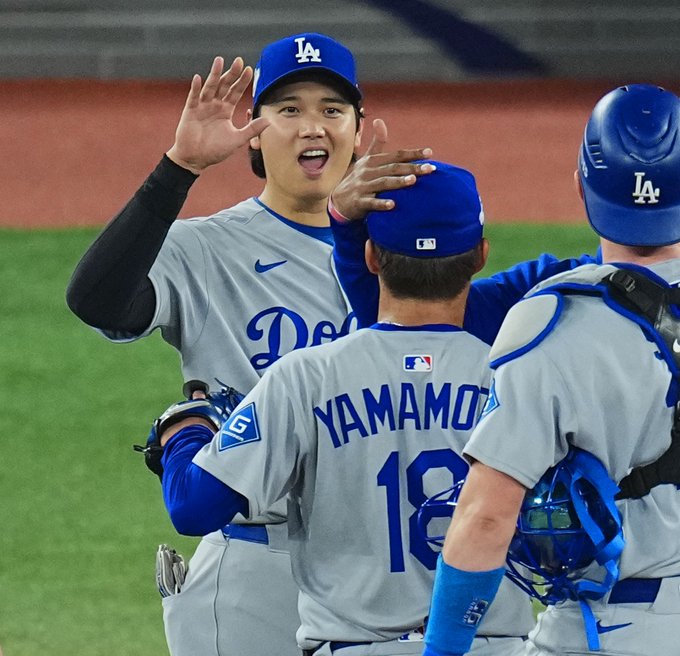 Shohei Ohtani raises both hands excitedly while touching Yoshinobu Yamamotos shoulder in celebration. Both wear Los Angeles Dodgers uniforms with blue hats and gray jerseys, Yamamoto numbered 18. A teammate in catchers gear stands nearby on green outfield grass under stadium lights.