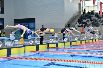 First image shows swimming pool with blue water and swimmers in action, Turkish and Palestinian flags hanging above, banner reading Özgür Filistin Free Palestine, audience seated in stands. Second image depicts group of men in suits and casual attire sitting on red-draped bench in front of empty seats with Turkish flags, clock and sign visible. Third image captures line of young athletes in yellow uniforms holding Turkish and Palestinian flags, walking along poolside in sports hall with banner of mayor. Fourth image features young female swimmers diving from starting blocks into pool, wearing swim caps and suits, lane markers and officials present.