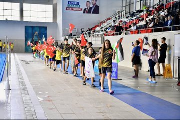 First image shows swimming pool with blue water and swimmers in action, Turkish and Palestinian flags hanging above, banner reading Özgür Filistin Free Palestine, audience seated in stands. Second image depicts group of men in suits and casual attire sitting on red-draped bench in front of empty seats with Turkish flags, clock and sign visible. Third image captures line of young athletes in yellow uniforms holding Turkish and Palestinian flags, walking along poolside in sports hall with banner of mayor. Fourth image features young female swimmers diving from starting blocks into pool, wearing swim caps and suits, lane markers and officials present.